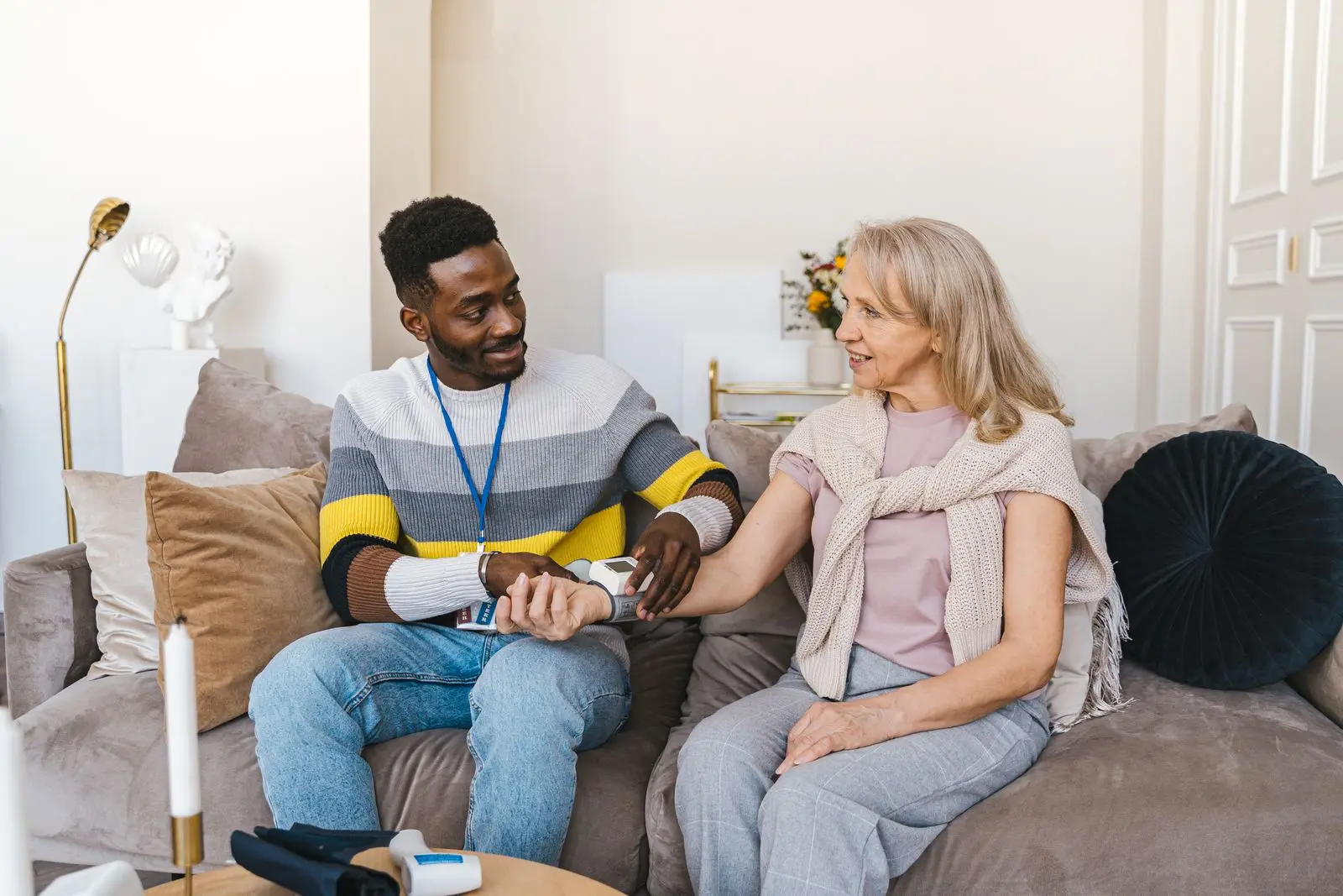 A smiling caregiver sitting with an older woman on her living room sofa, gently holding her wrist as they talk