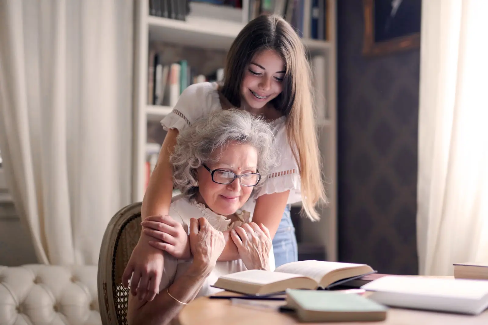 A granddaughter hugging her grandmother as she reads at a table