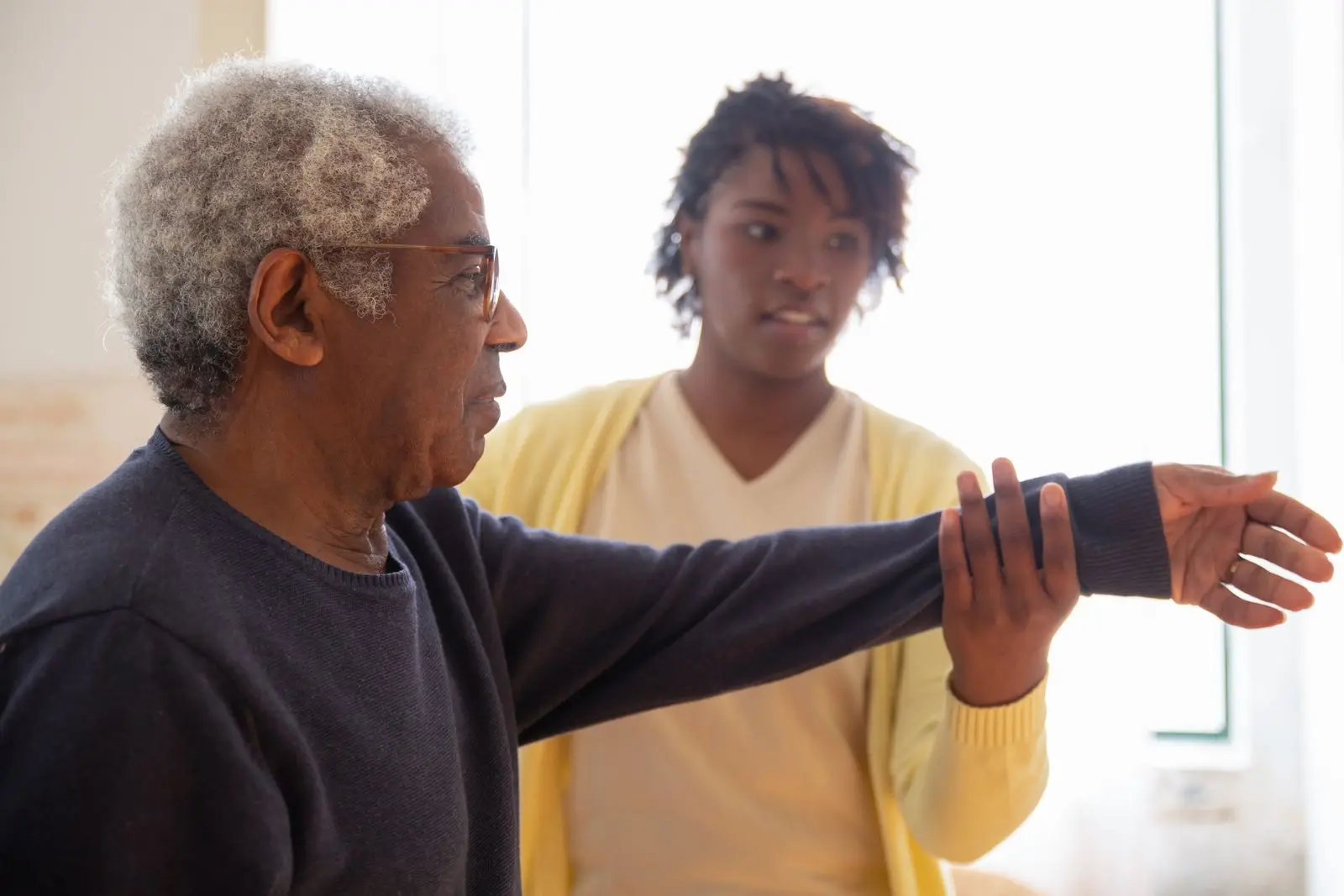 A caregiver supporting an older man's outstretched arm as he does a gentle stretch at home